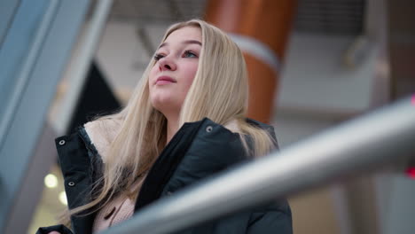 young lady with blonde hair and warm smile standing in shopping mall, she wears a winter jacket and looks content as she stands in a modern, stylish shopping center, surrounded by glass walls