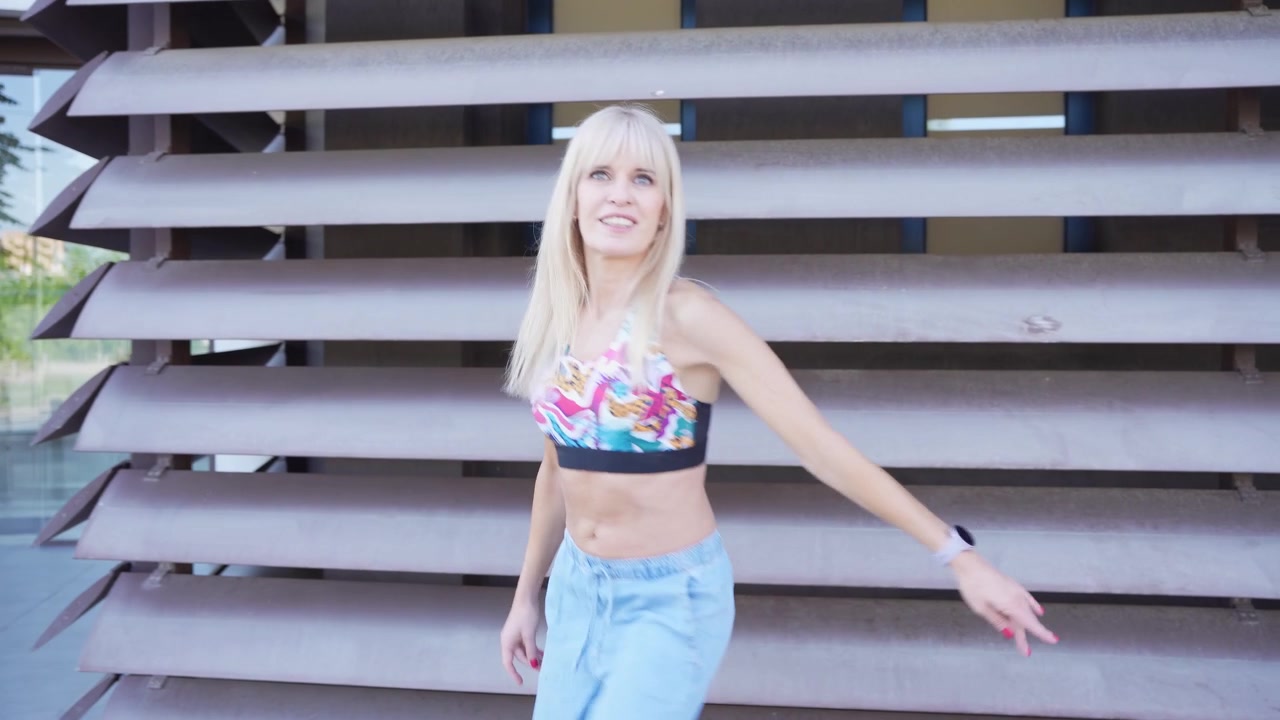 Una donna bionda in crop top e jeans balla con gioia all'aperto, mostrando uno stile di vita alla moda.