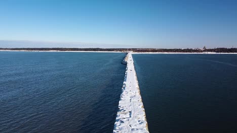 aerial view of snow and ice covered concrete pier in the calm baltic sea, port of liepaja on a sunny winter day, wide angle ascending drone shot