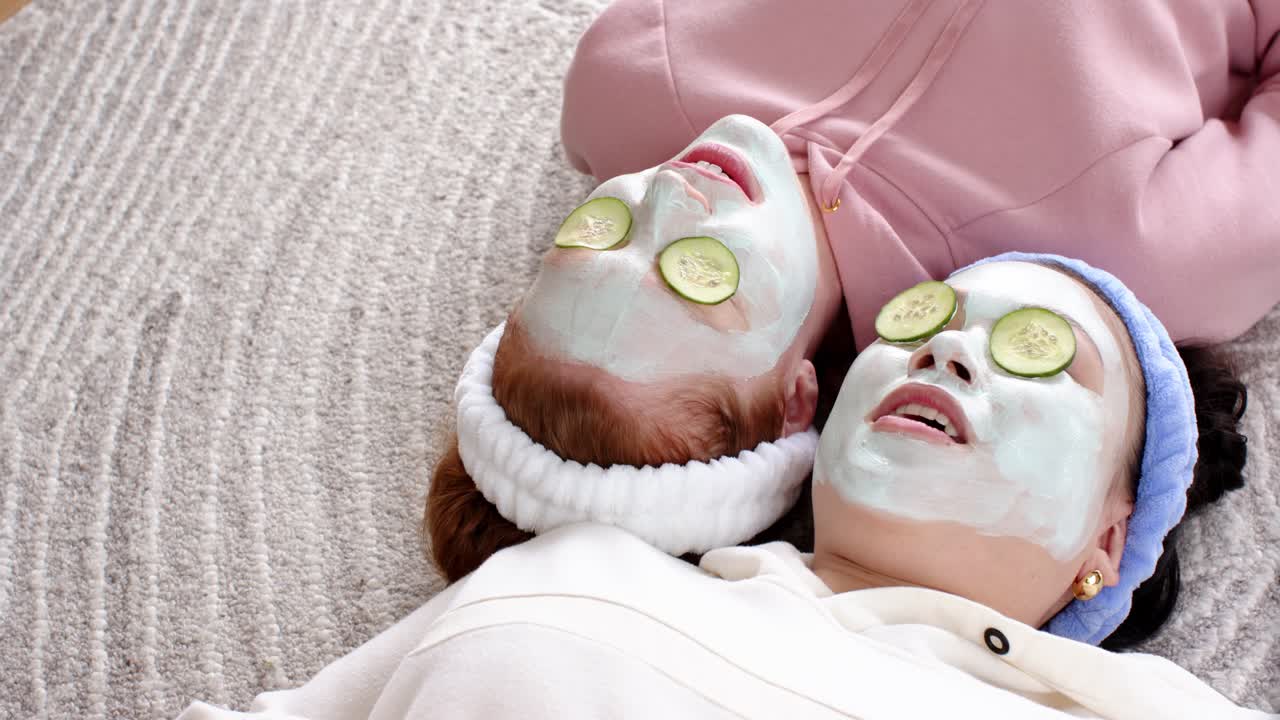 Hand entering frame causing two women lying on carpet moving mouths wearing clay masks, relaxing