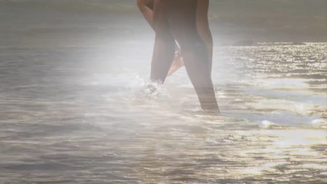 animation of legs of african american couple walking in sea at beach, over sun on sea