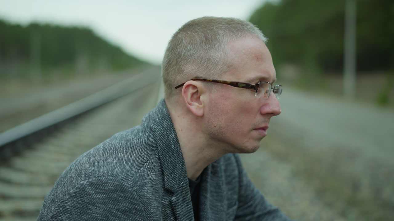 철도 트랙 에 앉아있는 안경을 입은 상하는 남자의 가까운 모습, 평온한 숲의 배경은 그의 사려 깊은 표정과 대조됩니다.