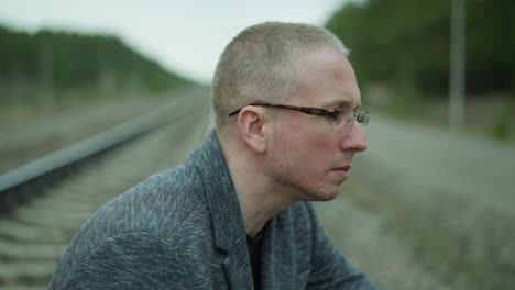 a close view of a contemplative man in glasses, looking away while sitting by a railway track, the serene forest background contrasts with his thoughtful expression