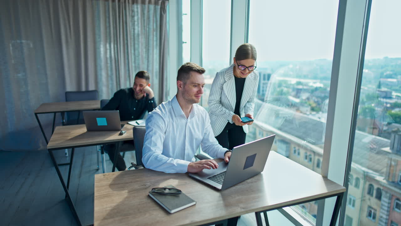 Teammates working in a modern light office. Man in white shirt works on his laptop and then notices something in the window.