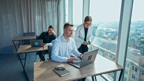 teammates working in a modern light office. man in white shirt works on his laptop and then notices something in the window.