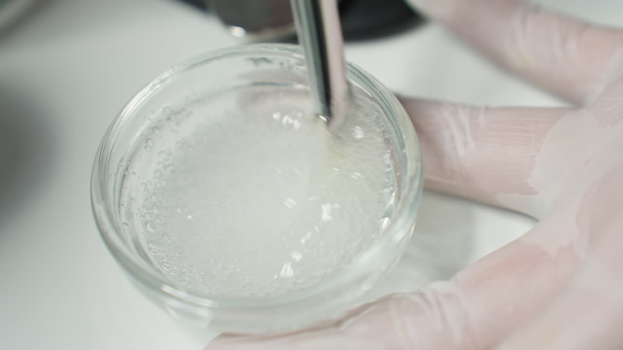 Practitioner hand in protective glove stirring gel face scrub with bubbles in small clear glass plate on white tray under soft light for professional facial treatment in modern spa environment