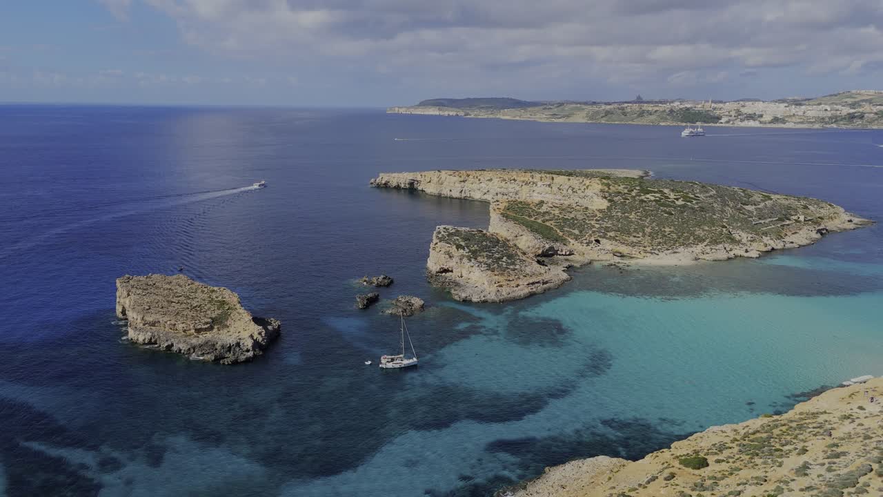 A high drone view over the Blue Lagoon shows Gozo, the open sea, part of Comino and Cominotto Beach with its cave, several yachts and a boat moving across calm turquoise water under sunny skies
