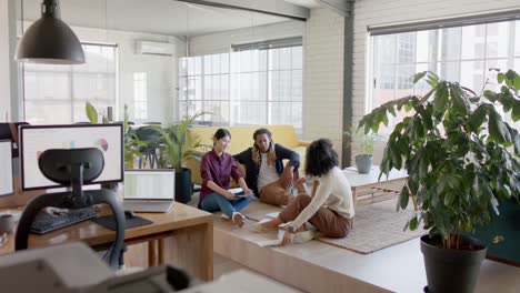 felices colegas creativos diversos usando tableta y discutiendo sentados en el piso de la oficina, cámara lenta