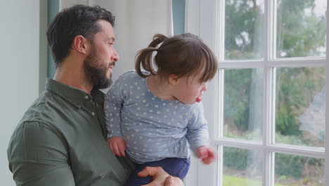 Padre-Amoroso-Sosteniendo-A-Su-Hija-Con-Síndrome-De-Down-En-Casa-Mirando-Juntos-Por-La-Ventana