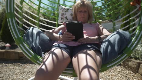 woman relaxes in comfortable garden swing chair reading digital ebook on warm sunny day