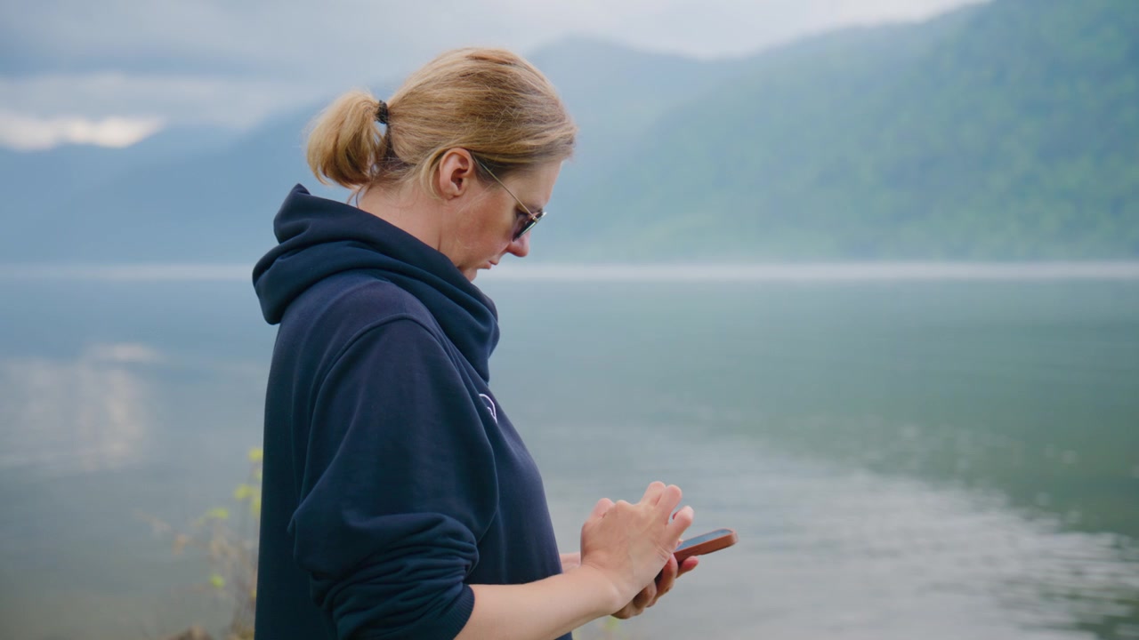 Kobieta stoi nad spokojnymi wodami, skupiona na telefonie. Ma na sobie ciemną bluzę z kapturem, włosy związane do tyłu. Mgła delikatnie unosi się nad jeziorem, a góry łagodnie wznoszą się w tle. Scena sprawia wrażenie spokojnej, cichej i refleksyjnej.
