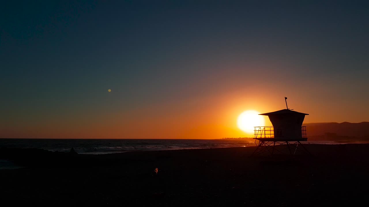 toma lenta de la puesta de sol lateral con la casa del salvavidas: torre que bloquea el sol mientras pasa por la playa estatal de san buenaventura en ventura, california, estados unidos