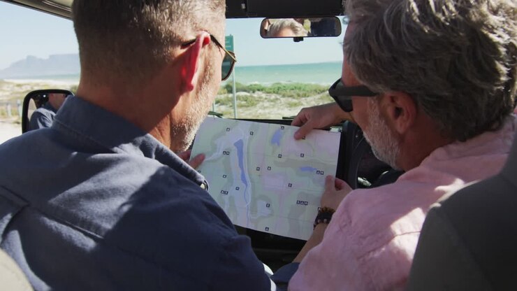 Felice coppia di uomini gay caucasici seduta in auto che legge una mappa e indica, in una giornata di sole al mare.