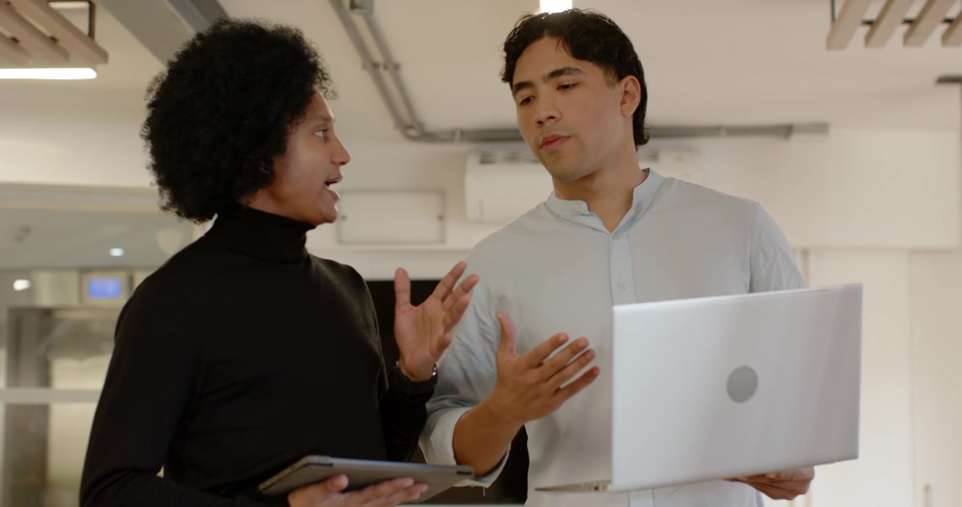 Diverse male coworkers, left holding tablet starting office talk, black top, right checking laptop