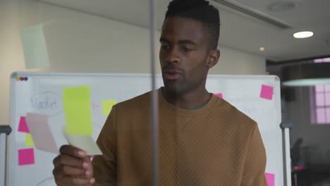 African-american-businessman-standing-behind-glass-wall-in-meeting-room-using-sticky-notes