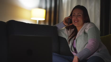 Smiling-Woman-Sitting-On-Sofa-At-Home-At-Night-Talking-On-Mobile-Phone-And-Watching-Movie-Or-Show-On-Laptop-1