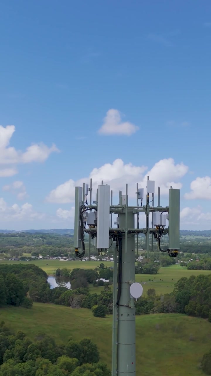 A tall communication mast stands centered against a blue sky and rolling green countryside.