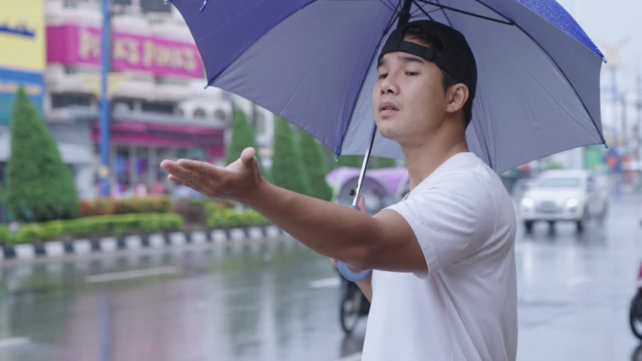 푸른 우산을 들고 길거리에 서 있는 젊은 아시아인, 방울, 비가 내리는 계절, 아지는 비, 병에 걸릴 위험, 에 혀있는 병, 호기심이 넘치는 손을 내밀어, 열대 태풍이 다가오고 있다.