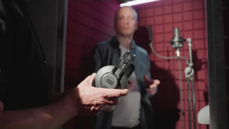 a focused scene in a recording studio where a man in a black shirt hands over headphones to another man in a blue long-sleeve shirt, set against a softly blurred red soundproof background