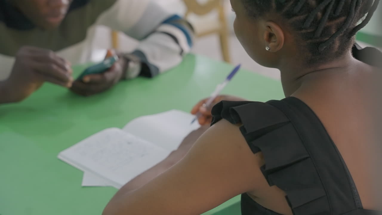 Black couple at table writing notes, woman scribbling in notebook while man points at phone, collaborative study date vibe, focused concentration, warm sunlight, green tabletop, attentive support