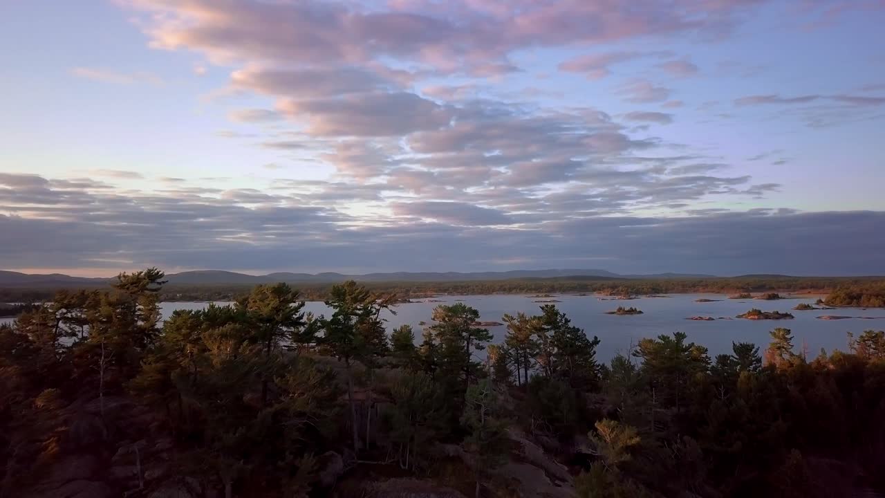 vuele sobre una colina de pinos rocosos para revelar pequeñas islas de granito, cielo azul y nubes al atardecer, una amplia plataforma aérea de drones en
