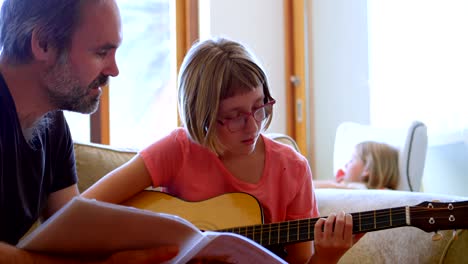 father helping her daughter to play guitar in living room 4k