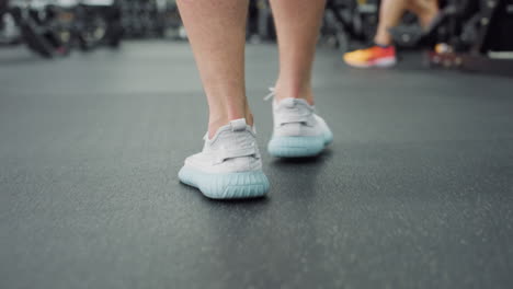 leg view of athlete wearing light blue sneakers moving across hall in fitness center, rubber flooring and blurred exercise machines around, highlighting motion, balance and active lifestyle