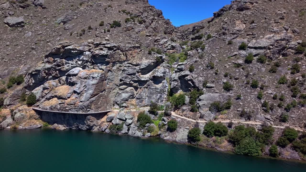 Aerial sideways drone view following the Dunstan Trail on a sunny day, showcasing rugged rocky hills and the Clutha River’s vivid blue waters in Central Otago, New Zealand