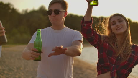 loving couple celebrate the end of the semester with beer and pop music on the beach with their friends. they are dancing on the open air party at sunset in summer evening.