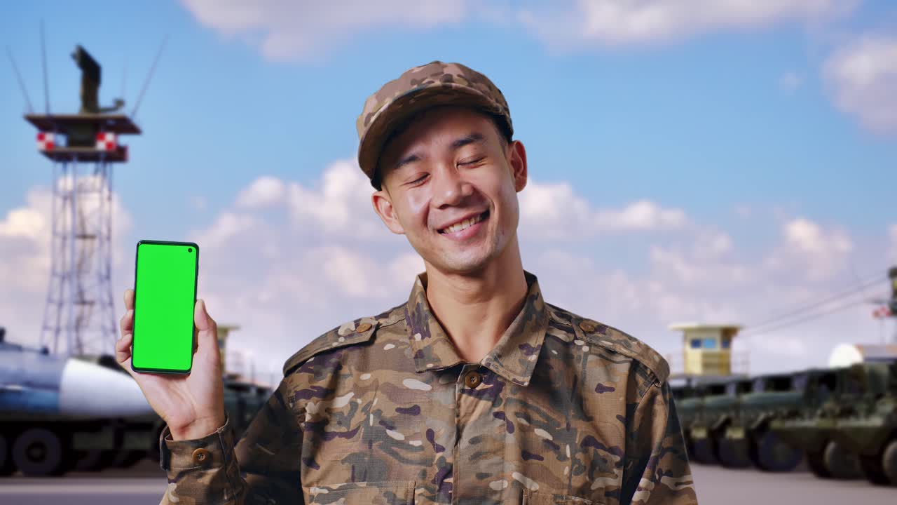 아시아 남성 군인이 미소 짓고 군 캠프에 서 있는 동안 카메라에 녹색 화면 스마트폰을 보여주고 있습니다.