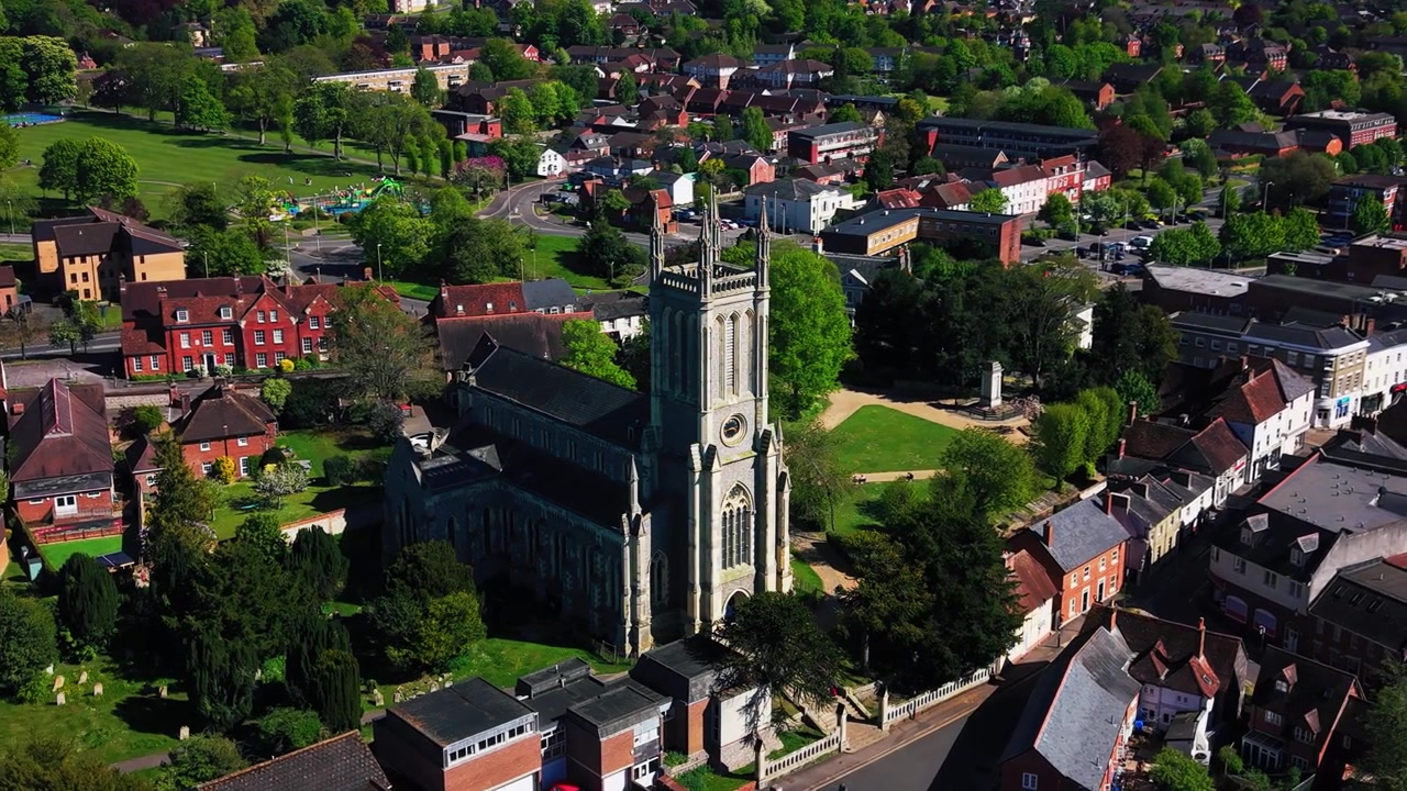Letecké záběry dronu míjející kostel sv. Marie v Andoveru, ukazující historickou kostelní věž, hřbitov, okolní ulice, střechy, zelené plochy a širší krajinu města Hampshire za slunečného dne.
