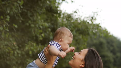 Madre-Alegre-Sosteniendo-A-Su-Hijo-Feliz-En-Las-Manos-Y-Riendo-En-El-Parque