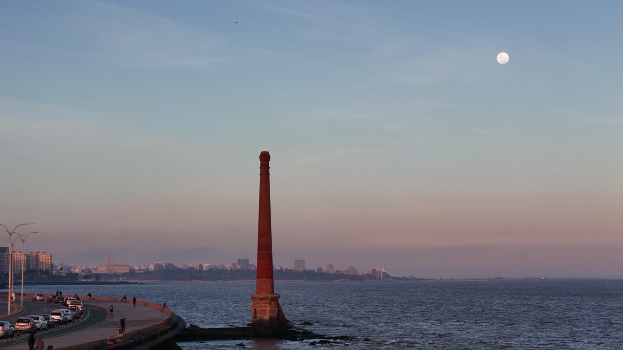 Aerial drone view over Montevideo’s Rambla Sur at sunset, featuring the city’s iconic chimney and the moon rising in the colorful evening sky