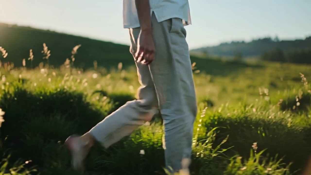 Lifting foot, barefoot adult walking through tall grasses at low sun, short-sleeved shirt swinging