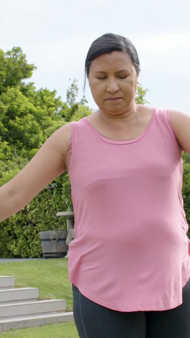 Video verticale: Una donna in equilibrio alza le braccia, facendo delle giravolte sul prato per mantenere l'equilibrio, con un top rosa e un tracker.