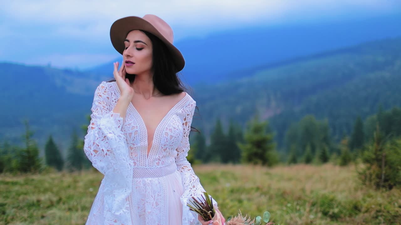 Romantic bride in white dress among nature. Portrait of a beautiful girl in wedding dress and hat on mountain landscape.