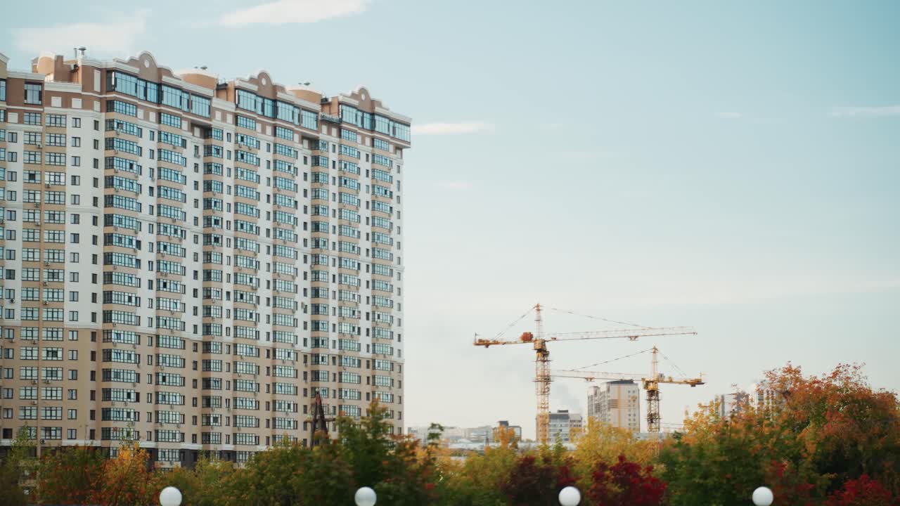 Highrise apartment under construction cranes rising, glass facade panels being installed, rooftop hoists in motion, site manager supervising facade installation, delivery van unloading materials