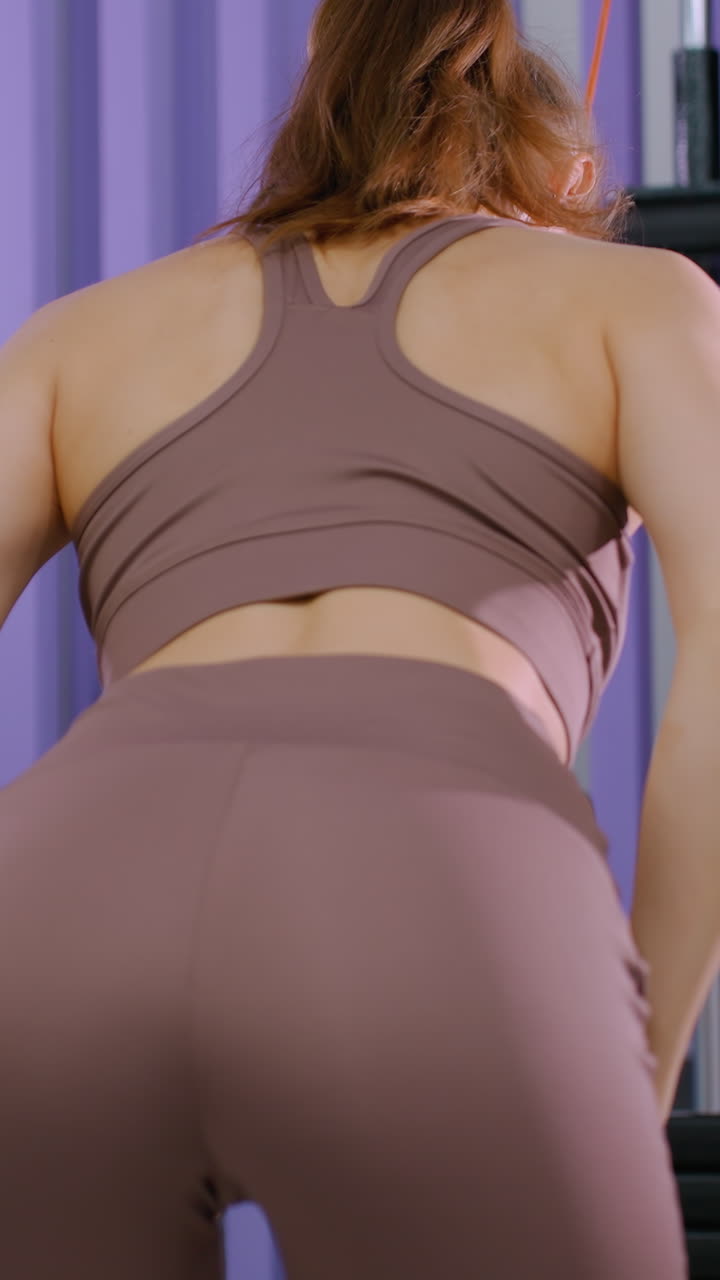 vista trasera de una mujer joven con ropa deportiva marrón realizando un tirón de pesas de cable en el gimnasio, involucrando los músculos del núcleo y la espalda, centrándose en el entrenamiento de fuerza, la rutina de fitness y el tonificación del cuerpo