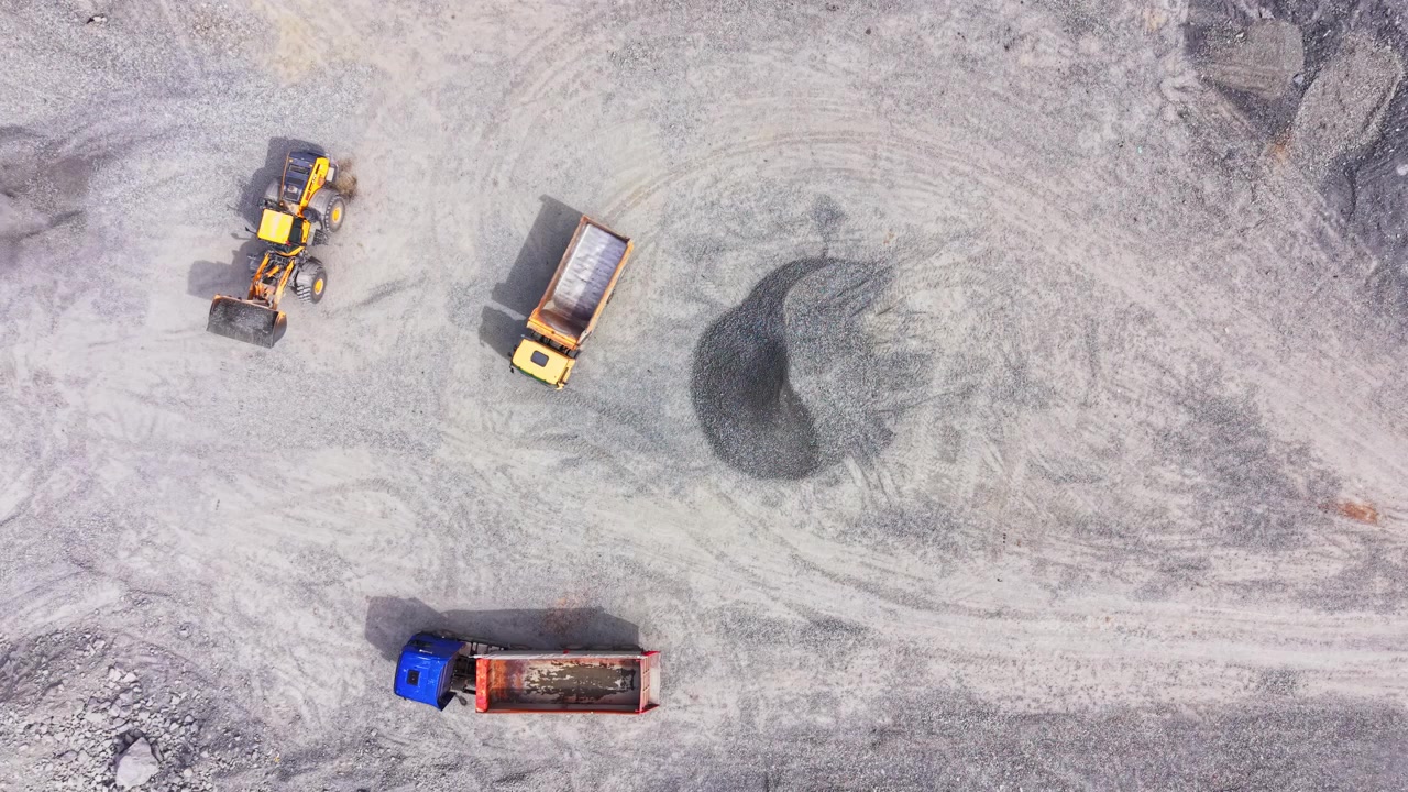 Drone view of quarry loaders and dump trucks converging. Overhead shot of gravel pile, four vehicles and crushed stone spread across the yard.