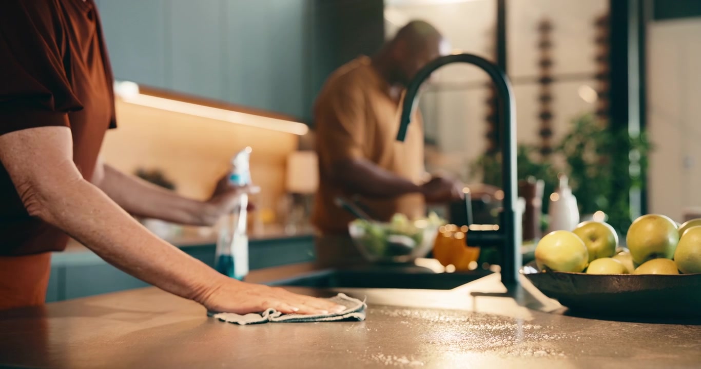 Woman Cleaning Kitchen Countertop While Man Prepares Food in Background