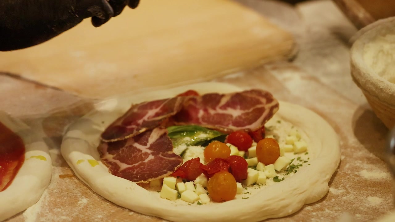 Mani guantate dispongono salumi sull'impasto della pizza con pomodori, formaggio e basilico. Primo piano.