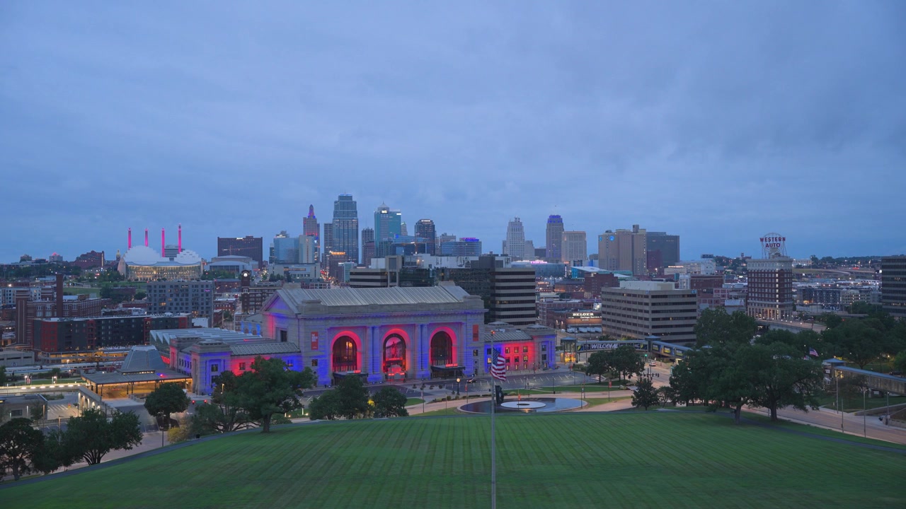 A locked-off aerial shot looks down over Union Station, its facade lit in red and purple, with the downtown Kansas City skyline glowing behind it. Overcast clouds hang low over the city as building an