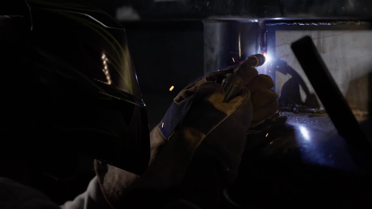 Close up of a metal worker's torch as it welds the side of a construction vehicle. Sparks fly in slow motion as sparks fly