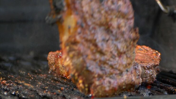 Delle pinze da carne girano una succosa costata di manzo (rib eye) quasi cotta su una griglia e la premono mentre le fiamme si alzano, al rallentatore.