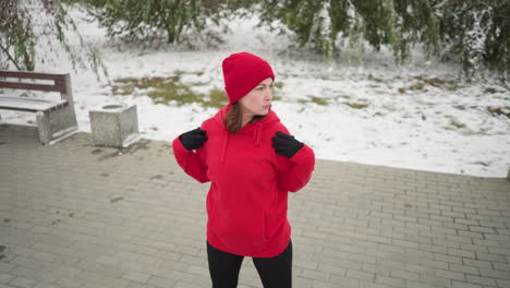 lady in red hoodie with hands placed on shoulders turns head slowly to left in snowy outdoor setting with frosted tree, bag on bench, and snow covering ground