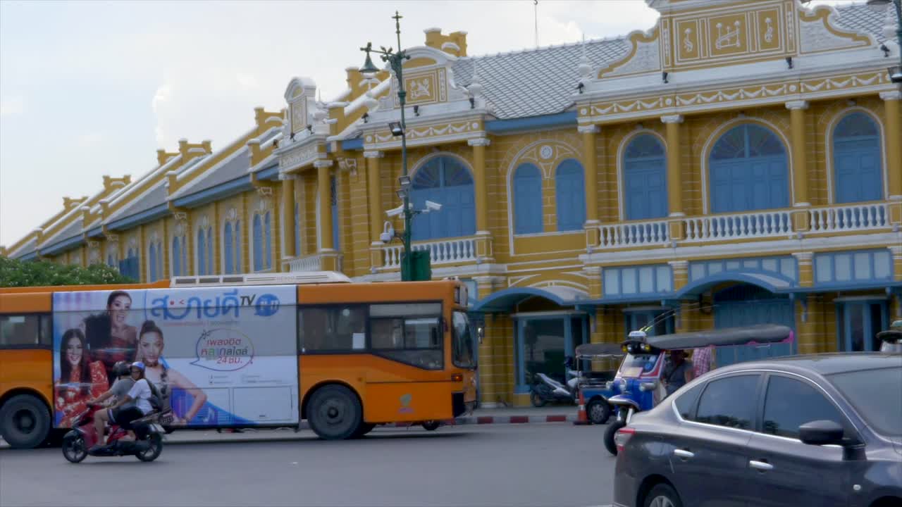 A crowded cross road with different vehicles passing by. Cars, Tuk Tuk, bus, scooters, taxi cross the scene in front of a colonialist and classic building with yellow walls and blue windows.