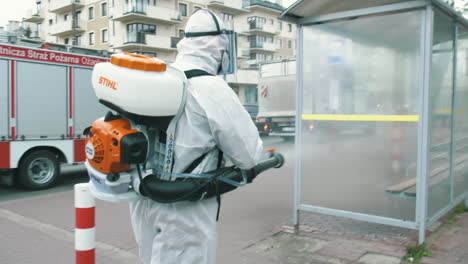man in the protective suit disinfecting bus stop with a spray gun to prevent coronavirus spread. spraying the gate