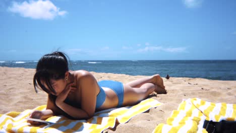 young black-haired woman reading on tablet device and sunbathing at picturesque beach