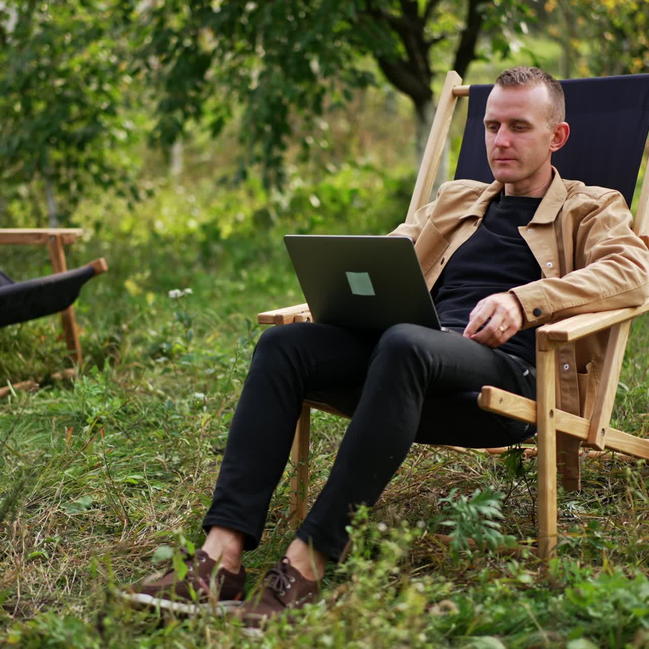 Calm relaxed freelancing man sitting in apple garden with laptop on his knees. Caucasian male working remotely online from nature