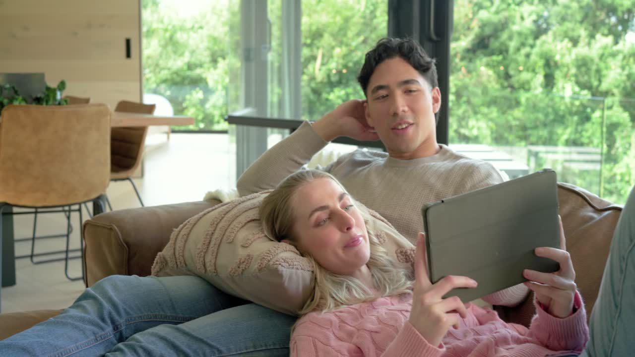 女性がタブレットをパートナーに向け、閲覧をガイドしながら、自宅の革製ソファでくつろぐカップル